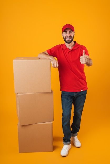 Portrait Of Handsome Delivery Man In Red Uniform Showing Thumb Up While Standing With Packaging Boxes Isolated Over Yellow Premium Photo